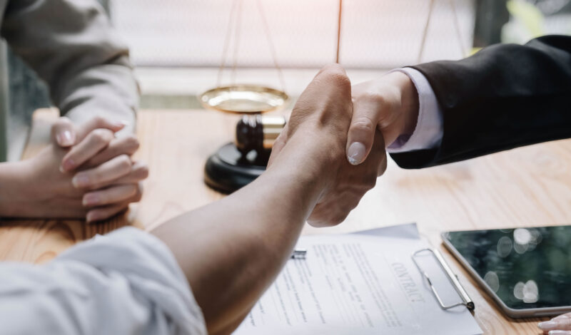 Close up of lawyer hand and man client shaking hand collaborate on working agreements with contract documents at the office.